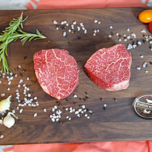 Two raw Wagyu tenderloin filets on a dark cutting board with rosemary, garlic and sea salt