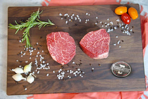 Two raw Wagyu tenderloin filets on a dark cutting board with rosemary, garlic and sea salt